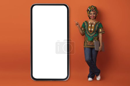 Téléchargez les photos : Joyeux positif attrayant jeune femme noire en costume africain avec ordinateur portable dans sa main pointant vers le gros téléphone avec écran blanc, montrant beau site Web ou une application, fond orange, espace de copie, maquette - en image libre de droit