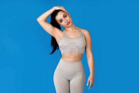 Photo for Sporty attractive well-fit young woman in tight beige sportswear stretching neck on blue studio background, athletic lady practicing yoga or pilates, have workout, copy space - Royalty Free Image
