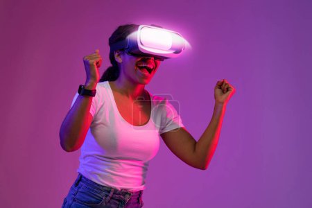 Téléchargez les photos : Femme noire ravie portant un casque VR célébrant le succès tout en explorant le cyberespace, femme afro-américaine émotionnelle criant d'excitation, debout dans la lumière au néon sur fond violet - en image libre de droit