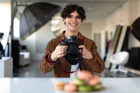 Téléchargez les photos : Jeune photographe alimentaire masculin utilisant un appareil photo numérique et prenant une photo du dessert, regardant et souriant à l'appareil photo. Photographe professionnel pendant le tournage de nourriture dans son studio - en image libre de droit