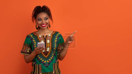 Téléchargez les photos : Sortir ensemble app. Positif sourire belle jeune femme afro-américaine en vêtements nationaux à l'aide d'un smartphone, pointant vers l'espace de copie pour la publicité, isolé sur fond de studio orange, panorama - en image libre de droit