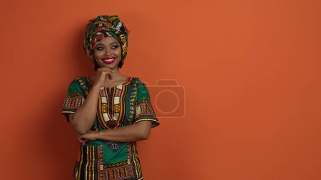 Foto de Buen trato. Impresionante mujer negra joven atractiva en traje africano brillante tradicional mirando el espacio de copia para el anuncio y sonriendo, tocando el pecho, aislado en fondo de estudio naranja, banner web - Imagen libre de derechos