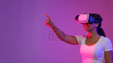 Photo for Virtual Experience. Smiling Black Woman In VR Headset Standing In Neon Light, Happy African American Female Touching Air With Hand While Playing Video Game, Enjoying Modern Technologies, Copy Space - Royalty Free Image