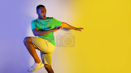 Photo for Athletic Black Male Exercising Looking Aside Over Blue And Yellow Neon Background. Studio Shot Of Sporty Guy Having Cardio Workout Jumping And Doing Elbow To Knee Crunches. Panorama, Copy Space - Royalty Free Image
