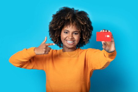 Foto de Feliz mujer afroamericana bastante milenaria con frenillos y en casual mostrando tarjeta bancaria roja en la cámara, sonriendo, recomendando el pago sin contacto, banca rápida fácil, fondo de estudio azul - Imagen libre de derechos