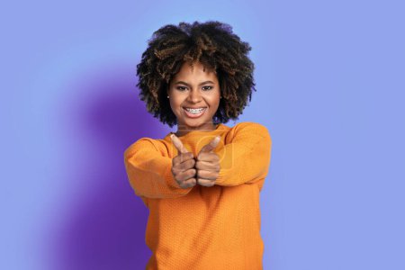 Téléchargez les photos : Positif heureux joyeux jeune femme afro-américaine en orange dans les cheveux touffus montrant pouces levés et souriant à la caméra sur fond de studio violet, recommandant un bon produit ou service - en image libre de droit