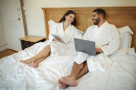 Téléchargez les photos : Couple joyeux en utilisant la tablette numérique et ordinateur portable fonctionnant en ligne et assis sur le lit à l'intérieur. Conjoints naviguant sur Internet sur les ordinateurs portant des peignoirs blancs dans la chambre moderne - en image libre de droit