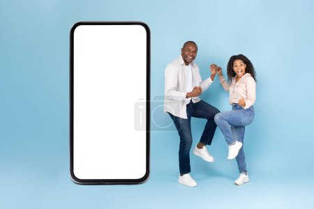 Photo for Excited black spouses gesturing yes in joy near huge cellphone with blank screen, advertising great mobile offer on blue studio background, full length, mockup - Royalty Free Image