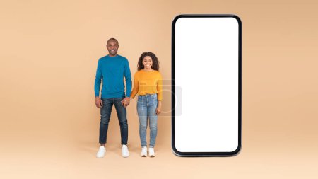 Photo for Happy african american spouses standing near huge cellphone with blank screen over peach background, advertising application or website, panorama. Full length, mockup - Royalty Free Image