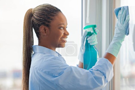 Photo for Closeup of happy smiling young black woman with long braided hair wearing workwear and rubber gloves cleaning windows, using spray and duster cloth, side view, maid service - Royalty Free Image