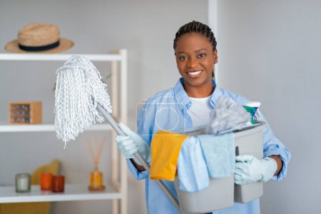 Photo for Professional cleaning service, house-keeping company. Cheerful pretty young black woman in workwear, rubber gloves holding cleaning supplies basket, apartment interior, copy space - Royalty Free Image