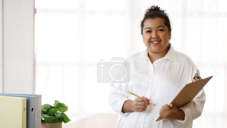 Foto de Mujer obesa atractiva de raza mixta joven en consejero de equipo formal que trabaja en la oficina, sosteniendo carpeta y pluma, sonriendo a la cámara, panorama con espacio para copiar. Empleo y ocupaciones - Imagen libre de derechos