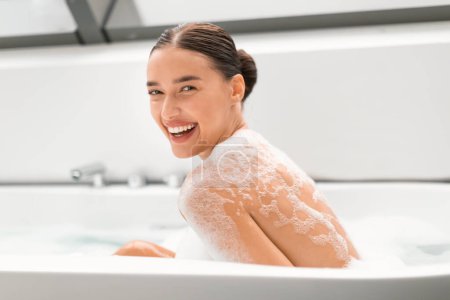 Téléchargez les photos : Jolie femme prenant bain avec de la mousse souriant regardant la caméra, se laver le corps et se détendre assis dans la baignoire dans la salle de bain moderne à l'intérieur. Rituels de beauté et concept choyer - en image libre de droit