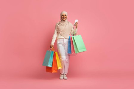 Photo for Smiling Muslim Woman In Hijab Walking With Smartphone And Shopping Bags In Hands, Arabic Shopaholic Lady Wearing Headscarf Enjoying App With Sales And Discount Offers, Pink Background, Copy Space - Royalty Free Image