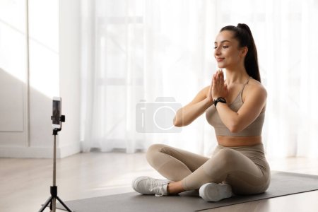 Foto de Clases de yoga en línea. Atractiva impresionante joven profesora de yoga tiene clase en línea, radiodifusión a través del teléfono en trípode mientras entrena en el gimnasio, tomándose de la mano en gesto namaste - Imagen libre de derechos