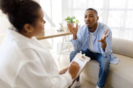 Photo for Psychotherapy. Anxious young black man in casual sitting on couch at clinic, looking at woman psychologist, sharing thoughts and gesturing, experiencing midlife crisis, have therapy session - Royalty Free Image