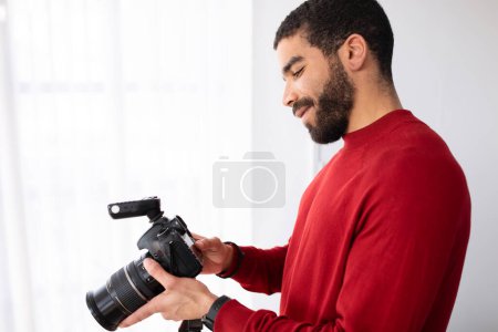 Téléchargez les photos : Photographe professionnel homme heureux beau barbu Moyen-Orient jeune homme en rouge avec appareil photo numérique en studio photo, vue latérale, espace de copie. Emploi et profession concept - en image libre de droit