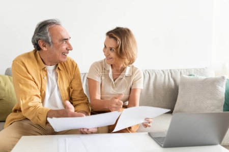 Photo for Senior couple holding documents reading paper bills, paying bank loan online, calculating taxes, planning family retirement money finances using laptop at home, free space - Royalty Free Image