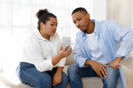 Foto de Mujer hispana bastante joven obesa mostrando su teléfono celular a su novio o esposo guapo chico negro, pareja de raza mixta sentada en el sofá en casa, revisando contenido extraño en línea - Imagen libre de derechos
