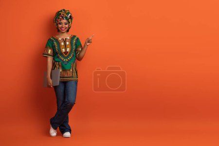 Photo for Positive cheerful pretty young black lady in african costume digital nomad posing with modern laptop on orange studio background, showing free space for ad. Freelance, remote job - Royalty Free Image