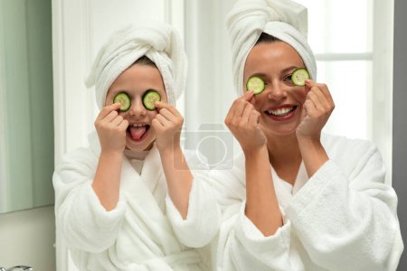 Photo for Cheerful european millennial woman and little girl in bathrobes and towel apply cucumber slices, have fun, take care of beauty and skin in white bathroom interior. Good morning, relationship - Royalty Free Image