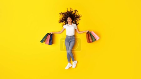 Photo for Shopping And Sales. Happy Female Shopaholic Holding Many Paper Shopper Bags Smiling To Camera Posing Over Yellow Studio Background. Seasonal Discounts Concept. Full Length Shot, Panorama - Royalty Free Image