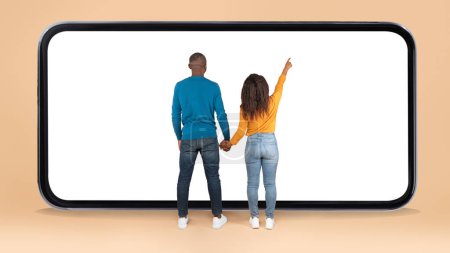 Photo for Back view of black spouses standing in front of huge smartphone with blank screen, pointing on display, standing on beige background, mockup, full length - Royalty Free Image