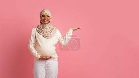 Foto de Bonita oferta. Sonriendo mujer musulmana embarazada demostrando algo en su palma vacía, alegre hermosa mujer de Oriente Medio en Hijab de pie sobre el fondo del estudio rosa, Panorama con espacio de copia - Imagen libre de derechos