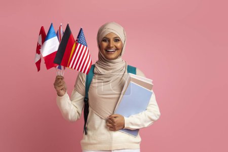 Téléchargez les photos : Portrait d'une étudiante musulmane souriante tenant des drapeaux internationaux et des cahiers d'exercices tout en se tenant au-dessus de l'arrière-plan rose en studio, heureuse femme islamique dans le hijab bénéficiant d'une éducation à l'étranger, Copier l'espace - en image libre de droit