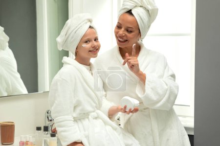 Téléchargez les photos : Heureuse femme millénaire européenne et adolescente en peignoirs et serviettes s'amusent, prennent soin de la beauté et de la peau, appliquent de la crème dans l'intérieur de la salle de bain blanche. Cosmétique, bonjour et relation - en image libre de droit