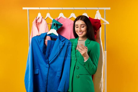 Photo for Mobile shopping. Contented woman shopaholic using cellphone and holding hanger with new jacket, standing on yellow background. Fashion and style application for smartphone - Royalty Free Image