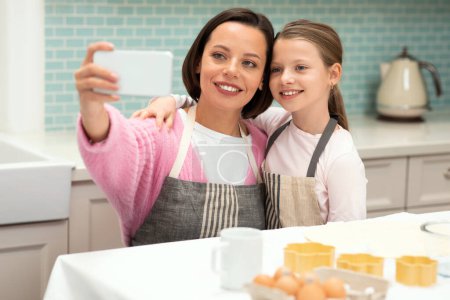 Photo for Happy european millennial woman taking selfie on smartphone with little girl in apron, preparing cookies in kitchen interior. App for blog, cooking homemade food, hobby in free time at home - Royalty Free Image