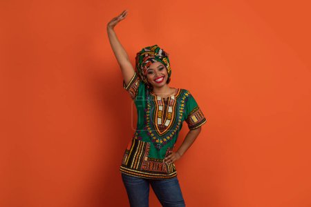 Foto de Mujer negra atractiva divertida positiva en traje africano colorido levantando la mano y sonriendo, señora bailando sobre fondo de estudio naranja. Diversión, alegría, concepto de emociones - Imagen libre de derechos