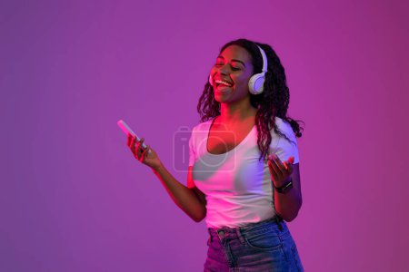 Téléchargez les photos : Femme noire insouciante portant des écouteurs sans fil écoutant de la musique sur Smartphone et chantant, Jeune afro-américaine joyeuse jouissant de chansons préférées, debout dans la lumière au néon sur fond violet - en image libre de droit