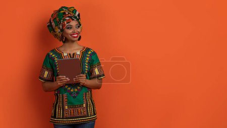 Foto de Alegre emocional atractiva joven africana mujer en traje nacional usuario femenino sosteniendo tableta digital y mirando espacio de copia sobre fondo de estudio naranja, web-banner - Imagen libre de derechos