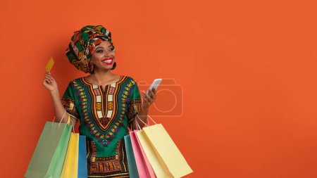 Téléchargez les photos : Jolie jeune femme africaine heureuse et rêveuse en costume national détenant des achats, téléphone portable et carte bancaire, regardant l'espace de copie et souriant, isolée sur orange, achats en ligne - en image libre de droit