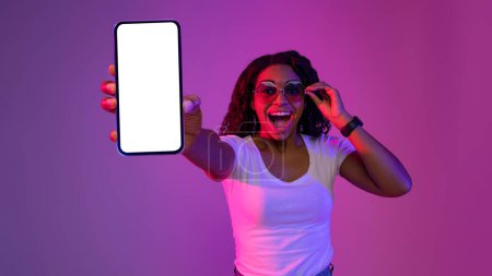Photo for Mobile Mockup. Excited Black Woman In Stylish Sunglasses Demonstrating Blank Smartphone At Camera, Amazed African American Lady Standing In Neon Light Over Purple Background, Copy Space - Royalty Free Image