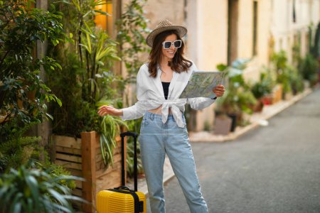 Photo for Cheerful millennial european woman tourist in hat and sunglasses with suitcase, map looking for travel route and hotel in city, outdoor. Journey alone, emotions from traveling on vacation - Royalty Free Image