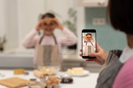 Photo for Happy european adult woman taking photo on smartphone screen of little girl in apron, making cookie dough, has fun on kitchen interior. Cooking homemade food, hobby at home, app for blog - Royalty Free Image