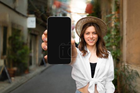 Photo for Glad pretty millennial european lady tourist in hat shows smartphone with empty screen, enjoy journey in city, outdoor, close up. Vacation travel, trip, blog, map app and website advice - Royalty Free Image