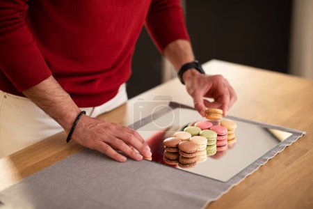 Photo for Cropped of male influencer or food blogger getting ready before photo shoot at home or photo studio, man hands setting sweets pastry colorful macarons on mirror surface, copy space - Royalty Free Image