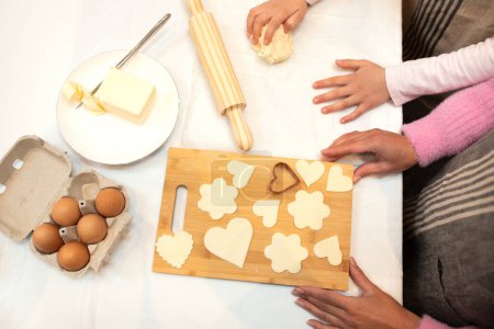 Foto de Niña europea y madre adulta hacen galletas de masa con rodillo de madera, harina y huevos en la mesa en la cocina, vista superior, recortado. Hobby y cocina, comida casera en casa - Imagen libre de derechos