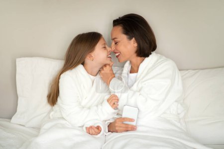 Photo for Cheerful european adult woman, little girl in bathrobes rub noses, sit on soft bed, enjoy rest together, free time, weekend and comfort in white bedroom interior. Good morning, relationship, love - Royalty Free Image