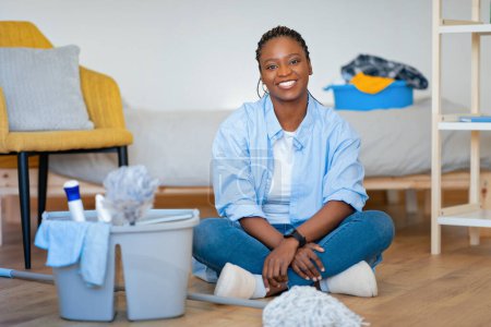 Téléchargez les photos : Positif heureux jolie jeune femme noire en vêtements de maison assis sur le sol à côté du panier avec des outils de nettoyage dans la chambre désordonnée, dame africaine au repos tout en faisant des tâches ménagères le week-end à la maison - en image libre de droit