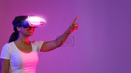 Foto de Mundo Virtual. Retrato de mujer negra emocionada usando auriculares VR tocando el aire mientras está de pie en luz de neón sobre fondo púrpura, sonriendo mujer afroamericana disfrutando de tecnologías modernas - Imagen libre de derechos
