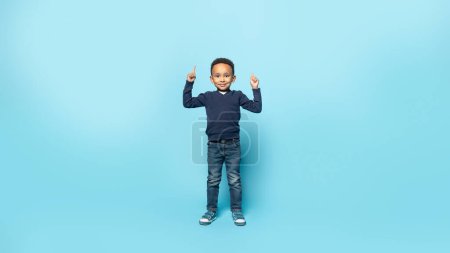 Téléchargez les photos : Levez les yeux. Adorable jeune garçon afro-américain pointant les doigts vers le haut à l'espace libre et souriant à la caméra, debout sur fond bleu studio, pleine longueur, panorama - en image libre de droit