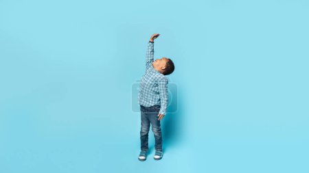 Photo for Little african american boy wishes he were taller, raising his hand to show how tall he wants to grow, child measuring his height, posing isolated on blue background, panorama, full length - Royalty Free Image