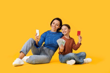 Photo for Online Shopping Concept. Happy Mother And Little Daughter Using Smartphone And Credit Card While Sitting On Floor Over Yellow Background, Family Buying Goods In Internet, Girl Holding Euro Cash - Royalty Free Image