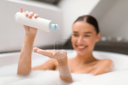 Téléchargez les photos : Lady Squeezing shampooing à portée de main prenant bain dans la salle de bain moderne à la maison, Focus sélectif sur la bouteille. Femme bénéficiant de soins du corps et des cheveux routine relaxant à l'intérieur. choyer et produits cosmétiques de bain - en image libre de droit