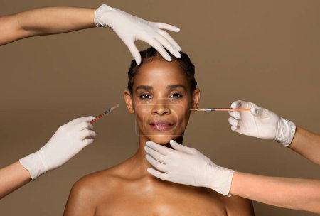 Foto de Atractiva mujer negra de mediana edad recibiendo inyección cosmética en su cara, cuatro manos de dos cosmetólogos. Inyección de ácido hialurónico para rejuvenecimiento facial - Imagen libre de derechos